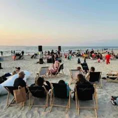 Wieczorne **krynica morska imprezy na plaży**. Ludzie siedzą na leżakach, słuchając muzyki przy zachodzącym słońcu.