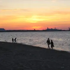 Zachód słońca nad morzem na plaży Stogi. Sylwetki ludzi spacerują brzegiem, w oddali widać statek i portowe dźwigi.