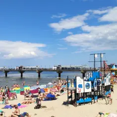 Tłum ludzi cieszy się słońcem i zabawą na plaży w Międzyzdrojach. W tle widać molo, a na piasku kolorowy plac zabaw.