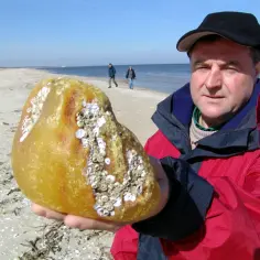 Mężczyzna trzyma duży kawałek bursztynu znalezionego nad morzem. Na plaży w tle widać dwie osoby.