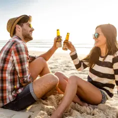 Para dwojga świętuje romantyczny weekend dla dwojga nad morzem, wznosząc tosty piwem na plaży.