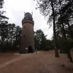 Czołpino atrakcje: ceglana wieża latarni morskiej pośród sosnowego lasu, z kilkoma osobami odpoczywającymi na ławkach w oddali.