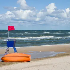 Czerwona flaga ostrzega przed niebezpieczeństwem. Zamknięte plaże Bałtyku, wywrócona łódź ratunkowa i wiejący wiatr.