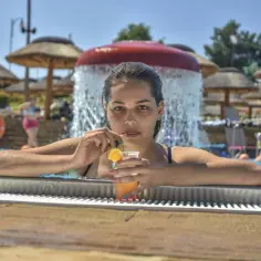 Kobieta z drinkiem w basenie, otoczona palmowymi parasolami i wodnym placem zabaw. To właśnie co oznacza all inclusive – beztroski relaks.