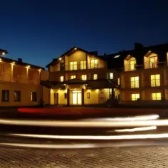 Elegancki hotel spa Rusałka nocą, z oświetlonymi oknami i balkonami. Smugi świateł samochodowych na brukowanym dziedzińcu.