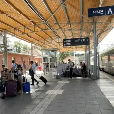 Ludzie z bagażami czekają na peronie. Znak "sektor A" i "peron 2" sugerują, że to stacja kolejowa, gdzie można dowiedzieć się, jak dojechać do Kołobrzegu pociągiem.