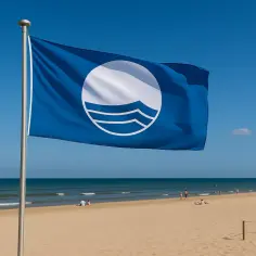 Biało niebieska flaga na plaży symbolizuje czystość wody i bezpieczeństwo. W tle widać spokojne morze i ludzi odpoczywających na piasku.