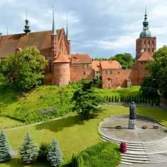 Zamek Frombork z katedrą i wieżą, otoczony zielenią. Na pierwszym planie pomnik na okrągłym placu.