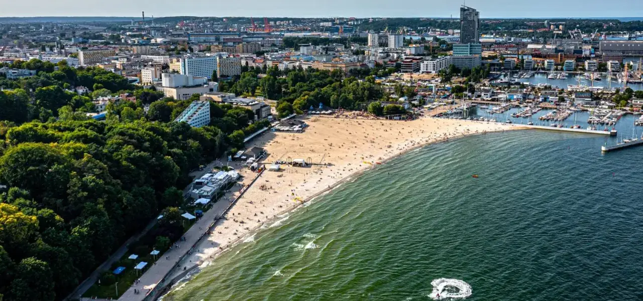 Słoneczny dzień na bulwarze nadmorskim w Gdyni. Plaża pełna ludzi, morze i marina pełna jachtów.