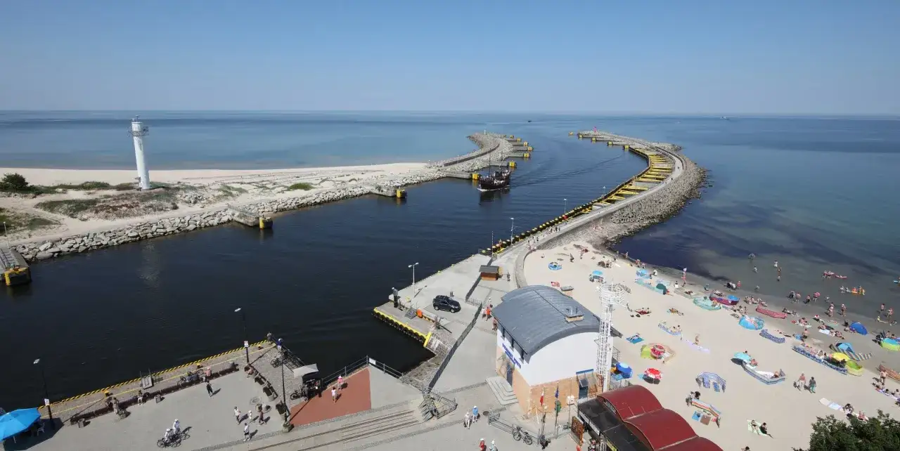 Słoneczny dzień w porcie Kołobrzeg. Statek wypływa z kanału, a plaża tętni życiem.