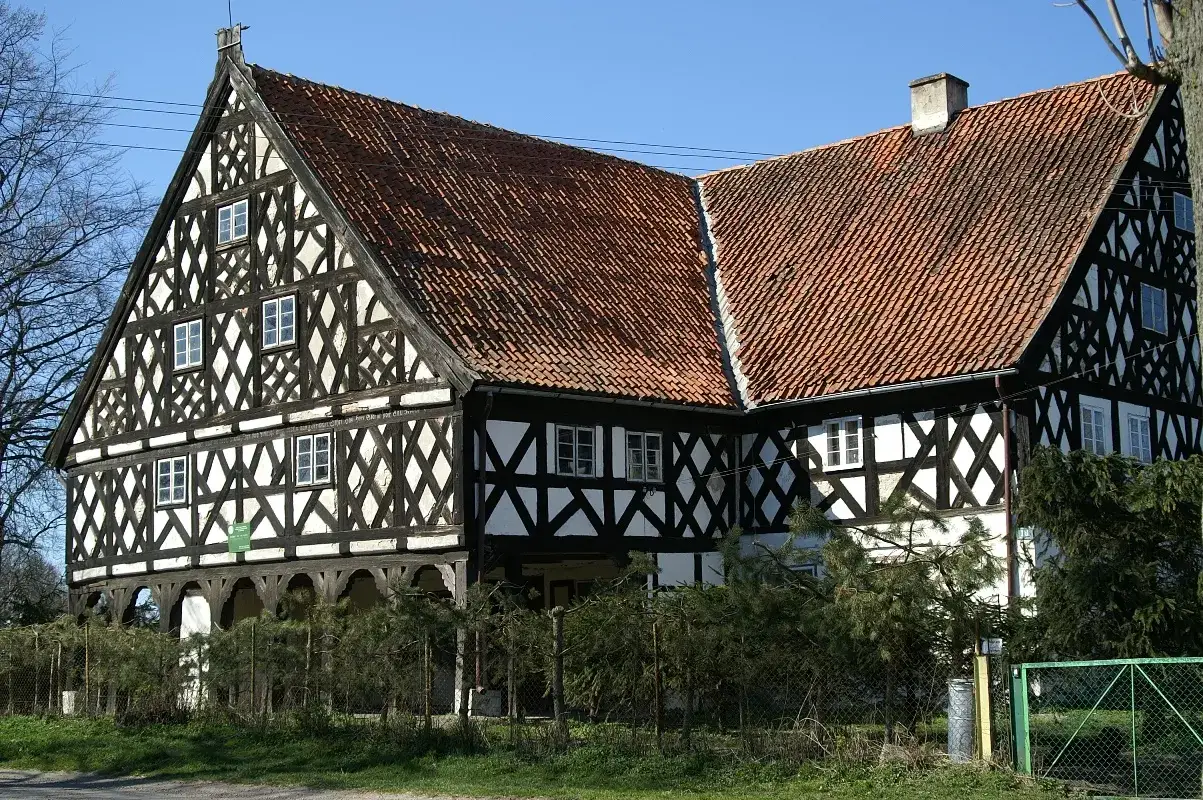 Charakterystyczny dom podcieniowy na Żuławach Wiślanych, z czarno-białą szachownicową fasadą i czerwoną dachówką.
