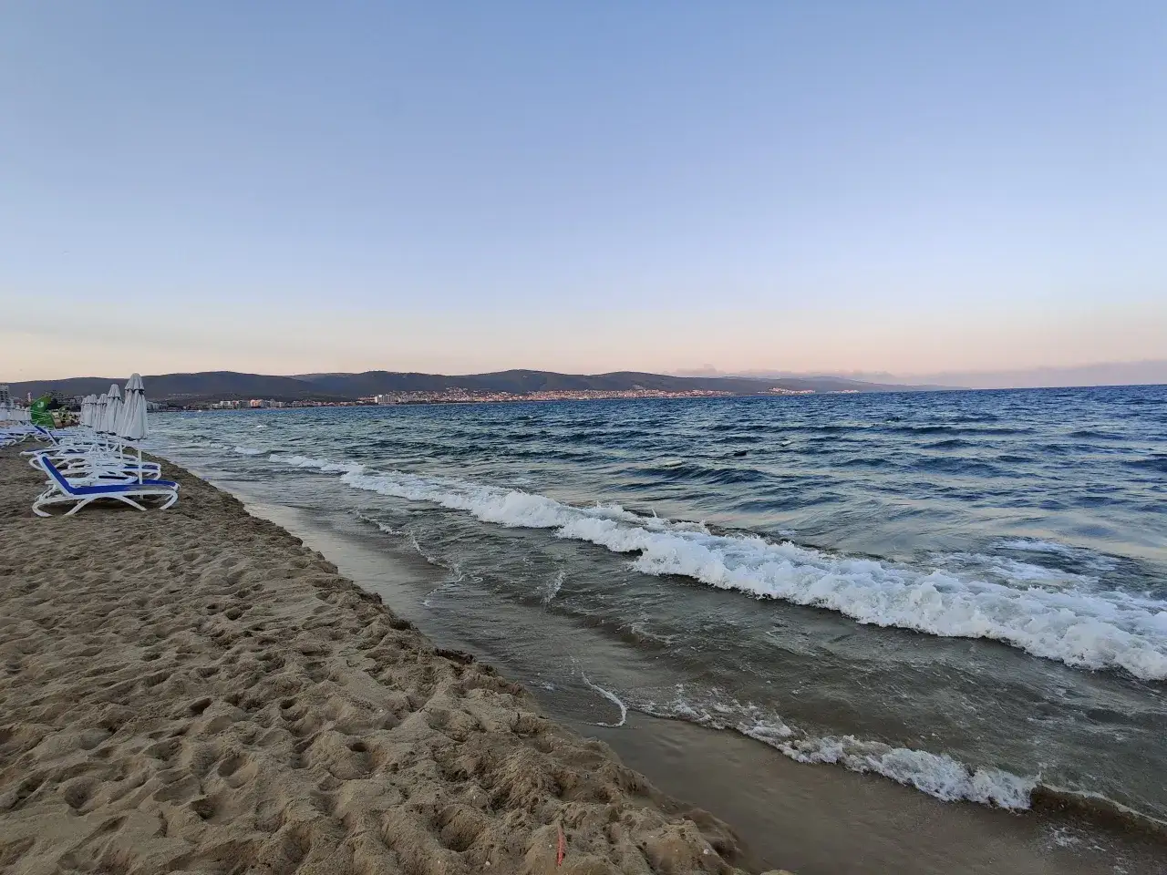 Piasek, leżaki i fale na plaży. Widok na miasto w oddali potwierdza, że Bułgaria ma dostęp do morza.