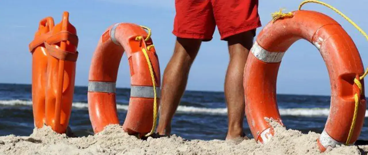 Ratownik wodny w czerwonych spodenkach stoi na plaży obok bojki ratowniczej i innych akcesoriów.