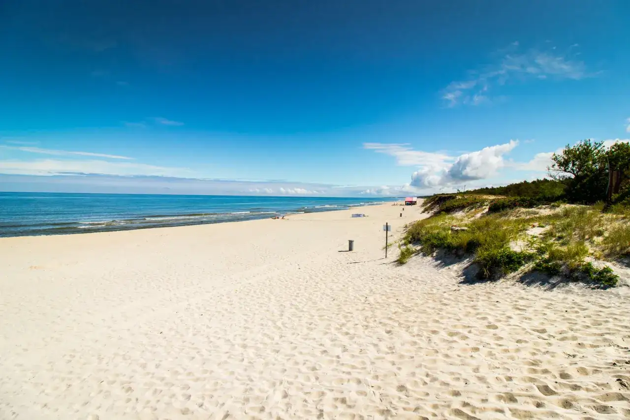 Piękna, piaszczysta stegna plaża z błękitnym niebem i spokojnym morzem. W oddali widać kilka osób i czerwony budynek ratownika.