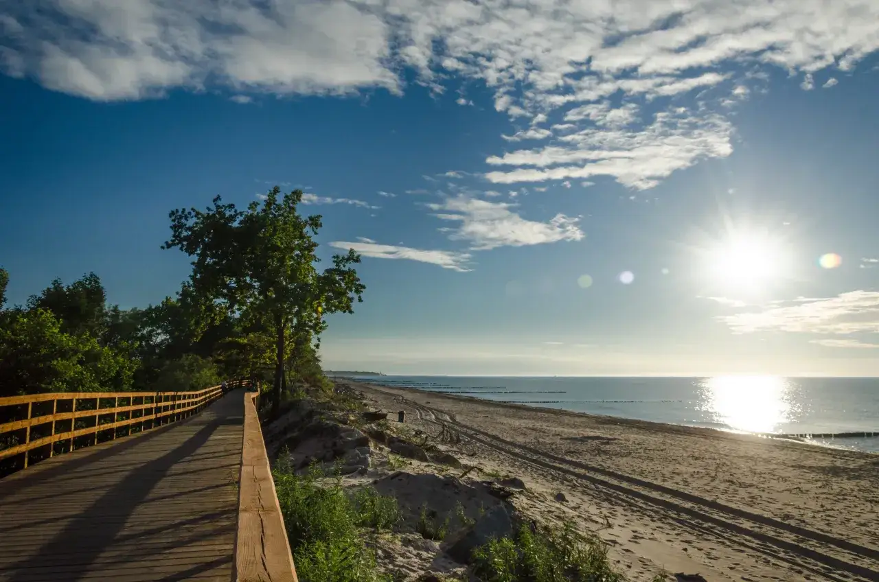 Drewniana ścieżka wzdłuż plaży, idealna na przejażdżkę rowerową Velo Baltica. Słońce odbija się w morzu, a chmury malują niebo.