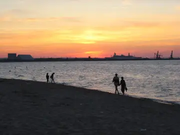 Zachód słońca nad morzem na plaży Stogi. Sylwetki ludzi spacerują brzegiem, w oddali widać statek i portowe dźwigi.