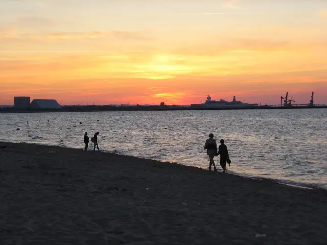 Zachód słońca nad morzem na plaży Stogi. Sylwetki ludzi spacerują brzegiem, w oddali widać statek i portowe dźwigi.
