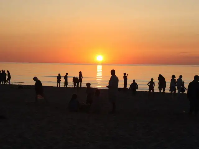 Zachód słońca w Sarbinowie. Ludzie spacerują po plaży, podziwiając pomarańczowe niebo i odbijające się w wodzie słońce.